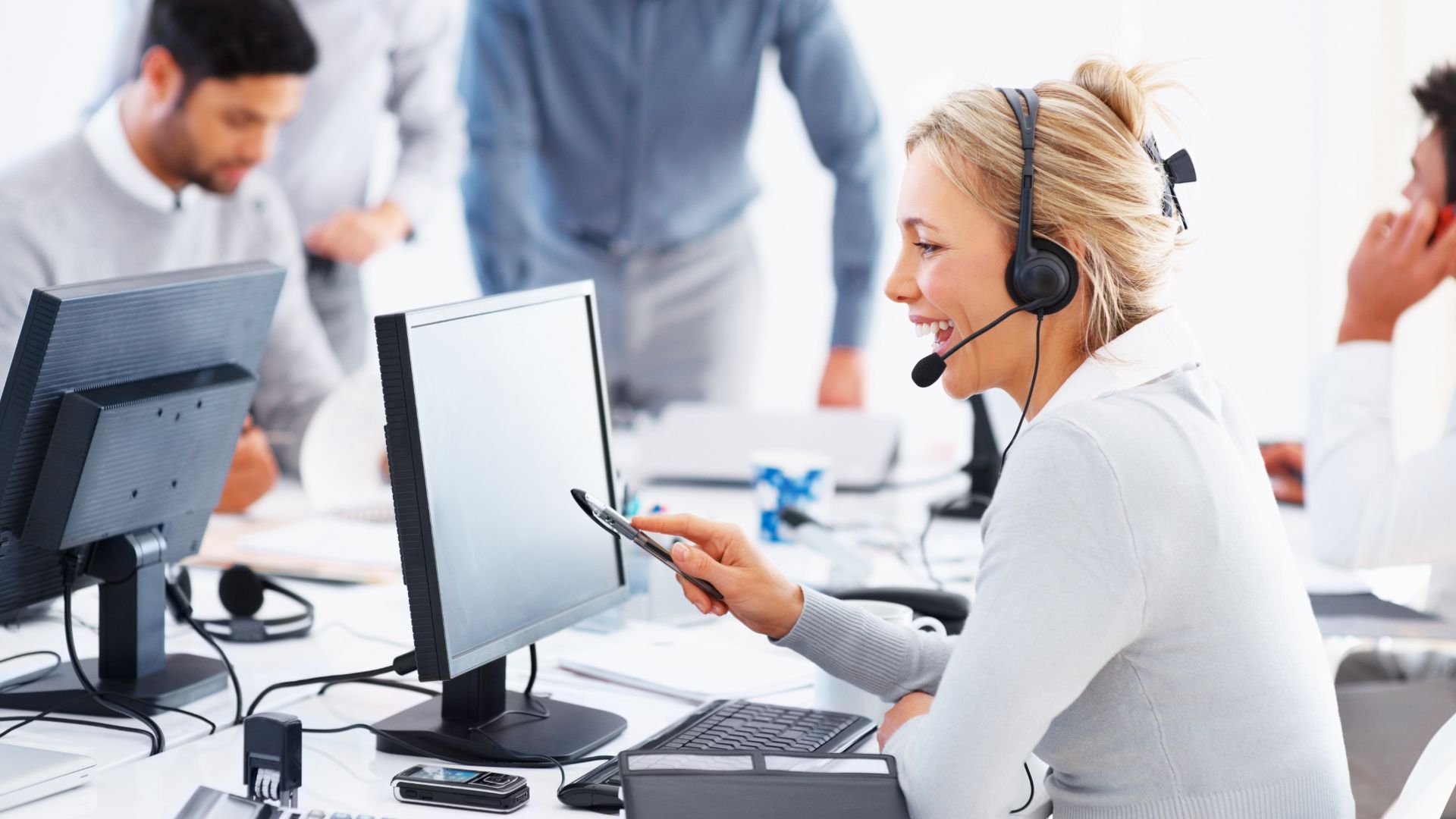 A woman on a phone call equipped with headphones and a computer and her busy colleagues in the background